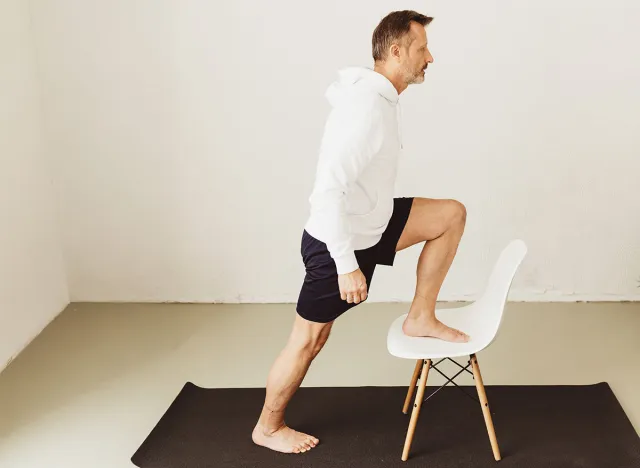 Middle-aged man doing step up stretch exercises using a chair during his daily workout at home during the Covid-19 pandemic