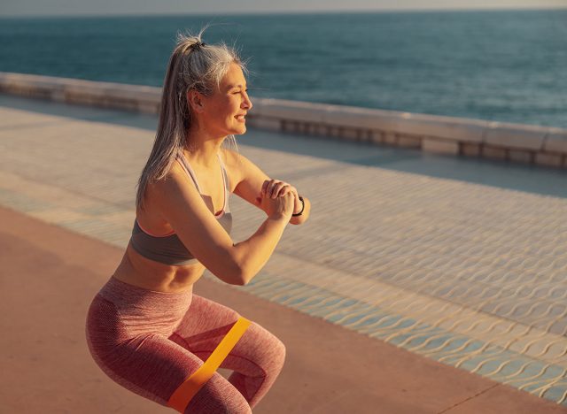 Adult woman training, performing squats with fitness band on sports street playground by the coast, shuffle