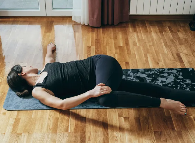 Practicing Reclined Spinal Twist Pose: Beautiful woman in sportswear doing spine rotation exercise on a gray exercise mat in the living room at home