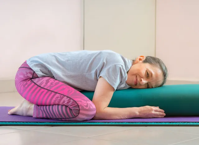Full length shot of energetic mature woman making exercising and practicing restorative yoga on mat and bolster at home because of social distancing, wearing sports clothes.