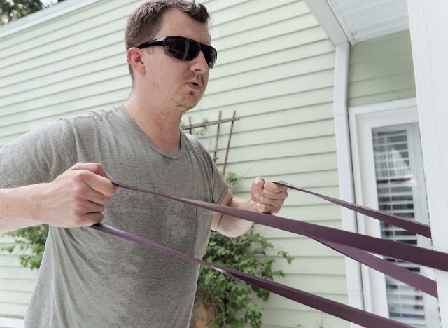 Man doing standing rows with a purple resistance band. This exercise is sometimes called "face pulls." He is doing an at-home workout.