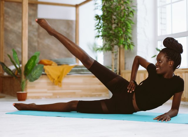 Beautiful athletic afro american woman lying on side and raising leg up selective focus, training on gym mat in decorated photo studio. Sports course, exercise technique, fitness, keeping body fit, lying leg circles 