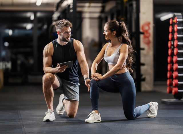 Personal trainer is showing a sportswoman how to do lunges correctly while kneeling in a gym.