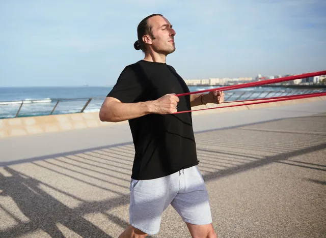Caucasian young adult athletic man in black t-shirt and gray sports shorts, exercising with resistance fitness band on Atlantic promenade, enjoying his bodyweight workout training on a warm sunny day