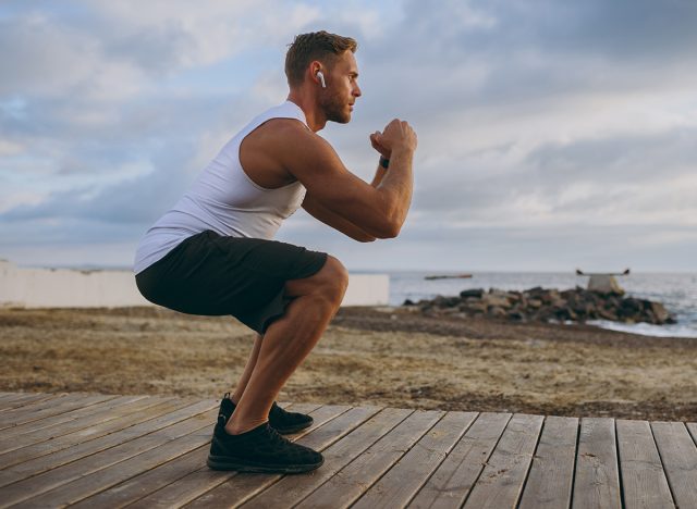 Full body young strong sporty athletic toned fit sportsman man wears sports clothes earphones do squats warm up train at sunrise sun dawn over sea beach outdoor on pier seaside in summer day morning, half squat pulse.