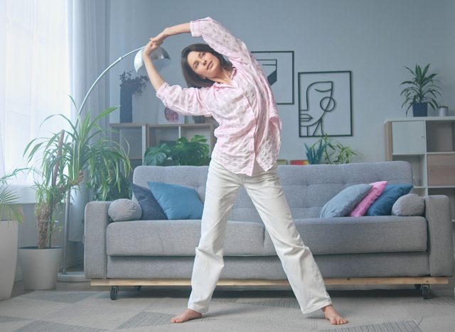 Woman is doing a side stretch in her living room, enjoying the natural light coming from the window. The room is decorated with modern furniture and houseplants