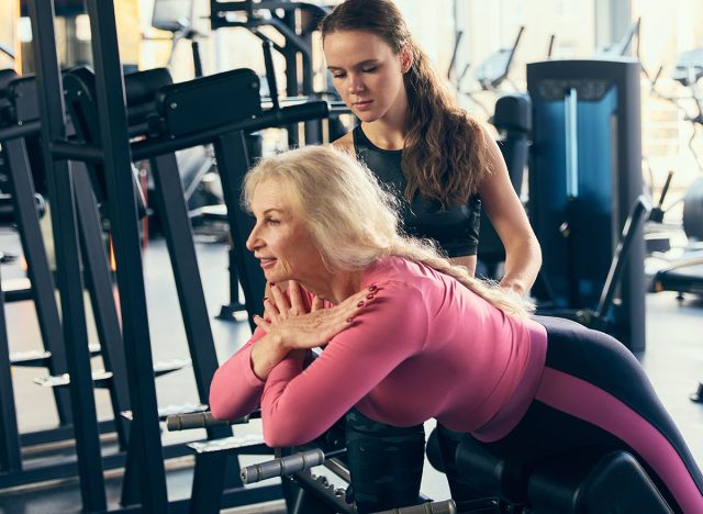 Fitness trainer assisting elderly woman with back extension exercises on machine in gym, focusing on spine health and strength. Concept of elderly people, sport, active lifestyle, health
