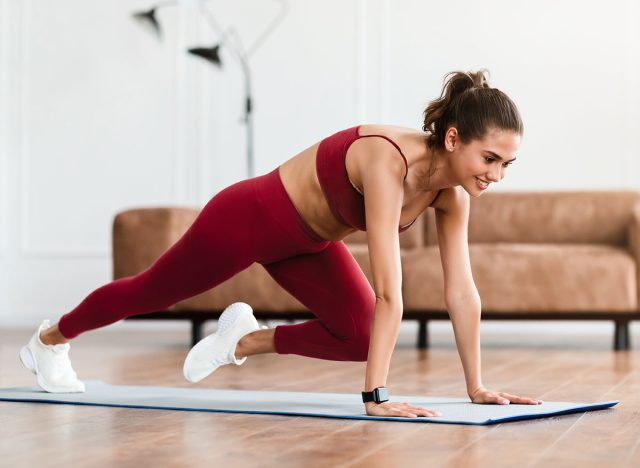 Smiling Sporty Woman Training At Home In Living Room In Front Of Laptop, Beautiful Female Doing Cross Body Mountain Climbers Exercise Looking At Screen Having Online Video Call With Trainer Instructor