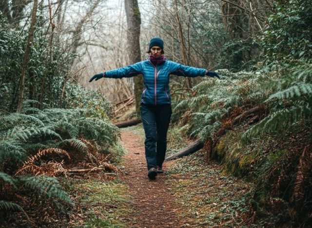 A serene outdoor scene with a person practicing tandem walking on a narrow trail in the forest, wearing hiking gear, surrounded by lush greenery, soft sunlight filtering through the trees, peaceful and inspiring atmosphere, winter season without snow