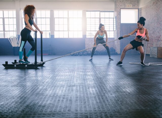 Shot of woman standing on sled with other pulling in gym. Three young females doing physical training at healthclub.