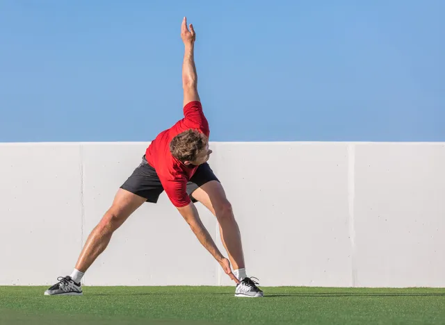 Windmill toe touch stretch dynamic stretching man standing training flexibility with back spinal twist exercise. Fitness athlete doing running warm up in summer park or home garden outdoors.