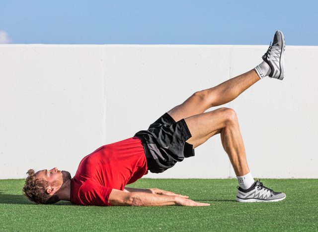 Fitness man doing bodyweight glute single leg floor bridge lift exercises. Fit athlete training glutes muscles with one-legged floor bridge butt raise in summer outdoor gym on grass.
