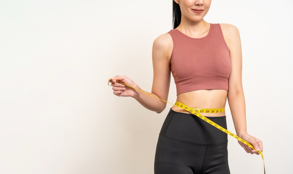 Close up woman measuring her waistline with measure tape. Weight loss diet session. Woman in sportswear slim fit and firm body shape. Care about eating and workout exercise.