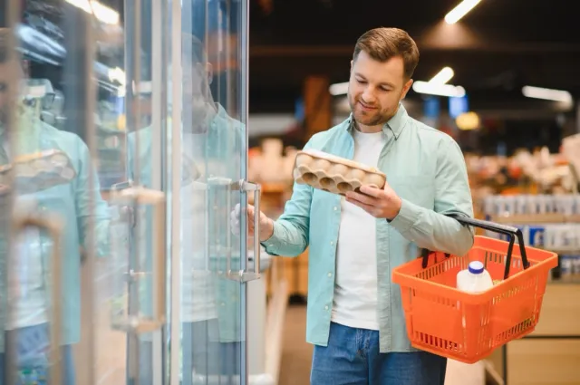 Man selecting fresh eggs from refrigerated section in grocery store, holding shopping basket with milk, demonstrating careful food choices