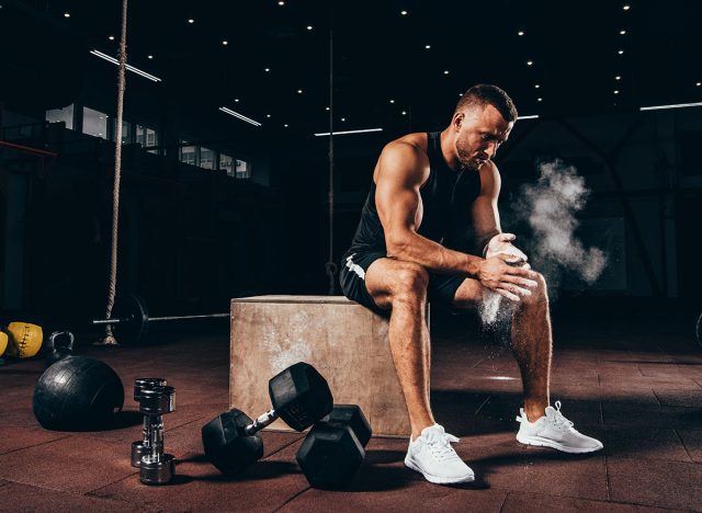 handsome athletic man sitting on cube with gym equipment around and clapping hands with talc before workout