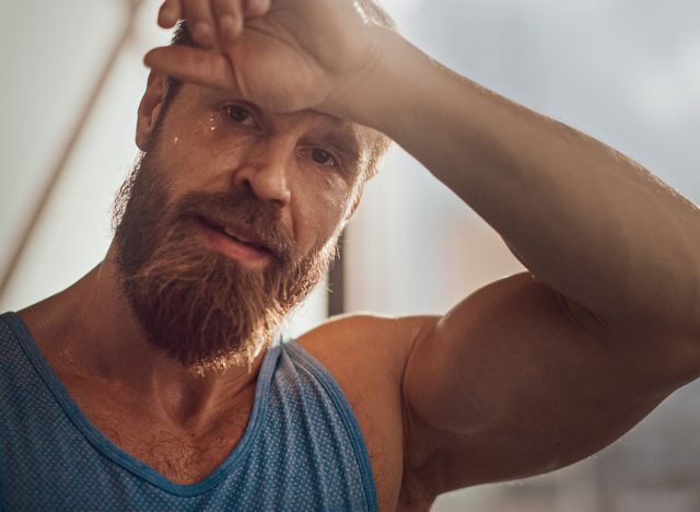 Portrait of Strong Bearded Male Athlete Wearing Sleeveless Shirt Wipes Sweat From His Forehead with His Muscular Hand. Handsome Man after Hardcore Exercise and Training. Man Gets Job Done