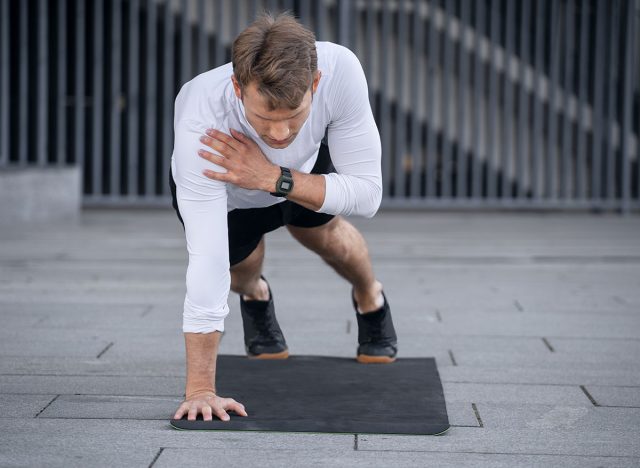 Full length view of young sportsman doing shoulder tap in push or press ups exercise, standing in plank position. Athlete with smartwatch training outdoors, warm up body, working out on yoga mat