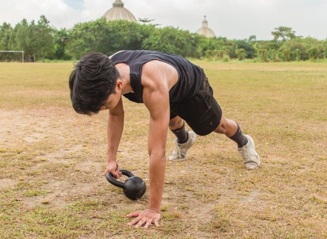 A young man does a set of Kettlebell plank drags. Advanced core and oblique workout. Training at an outdoor field.
