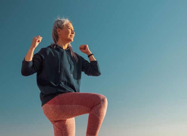 Active female in pink leggins and blue hoodie exercising , lifting one knee up and looking ahead on sunny day