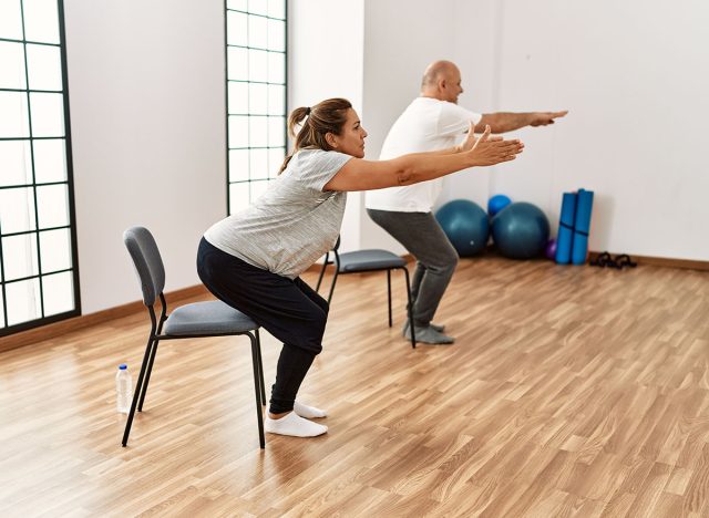 Middle age hispanic couple stretching using chair at sport center.