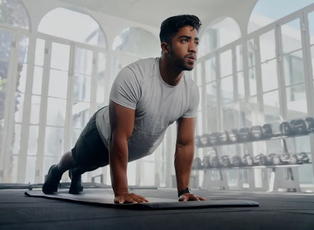 Young multiracial man in sportswear in plank position while doing push-ups on exercise mat at gym