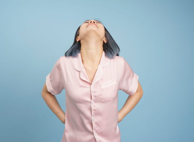 A young woman wearing pink pajamas stands confidently against a blue background. She has her hands on her hips, tilting her head back, exuding a sense of empowerment and self-assuredness.