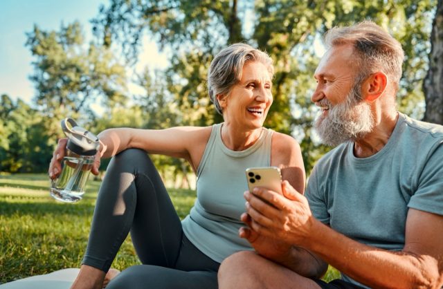 Gadget for recreation after sport. Cheerful couple with grey hair relaxing at park after workout and surfing internet on mobile. Beautiful woman holding water bottle while bearded man using cellphone.