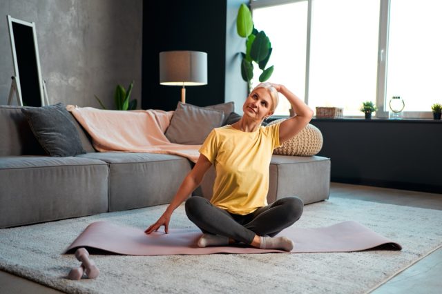 Sports and health care. Attractive mature woman doing neck exercise, exercising at home on a mat, sofa, big windows and decor in the background.