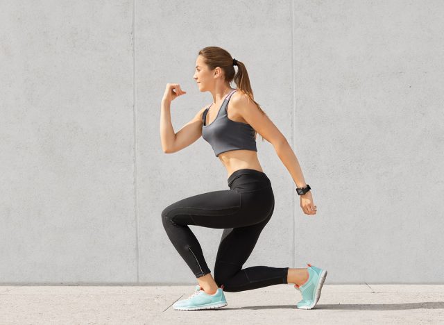 Indoor shot of self determined woman with pony tail, stands on one knee, warms up before cardio training, wears tanktop, leggings, sneakes, goes in for sport regularly, isolated over grey background