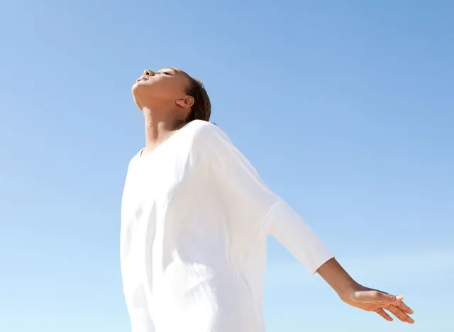 Young teenage girl breathing fresh air while standing by the sea with her arms outstretched and with her head leaning back against a blue sky.