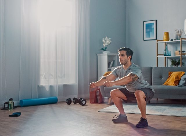 Muscular Athletic Fit Man in T-shirt and Shorts is Doing Squat Exercises at Home in His Spacious and Bright Living Room with Minimalistic Interior.