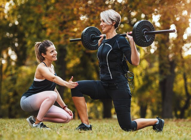 A mature woman is doing lunges exercises with personal trainer in the park, dressed in a black suit with an EMS electronic simulator to stimulate her muscles.