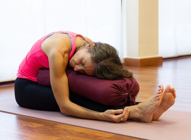 Middle aged woman on pink tank top in paschimottanasana pose resting chest on bolster at bright studio. Female yogi on seated forward bend asana. Therapeutic exercise concept