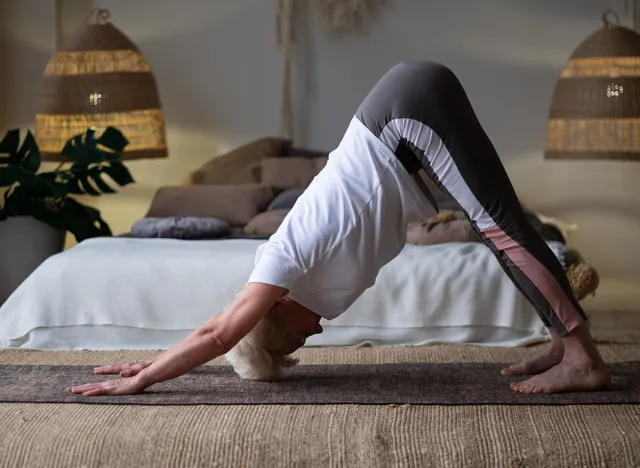 Senior woman working out at home, doing yoga exercise in the room downward facing dog pose, adho mukha svanasana fron sun salutation pose.