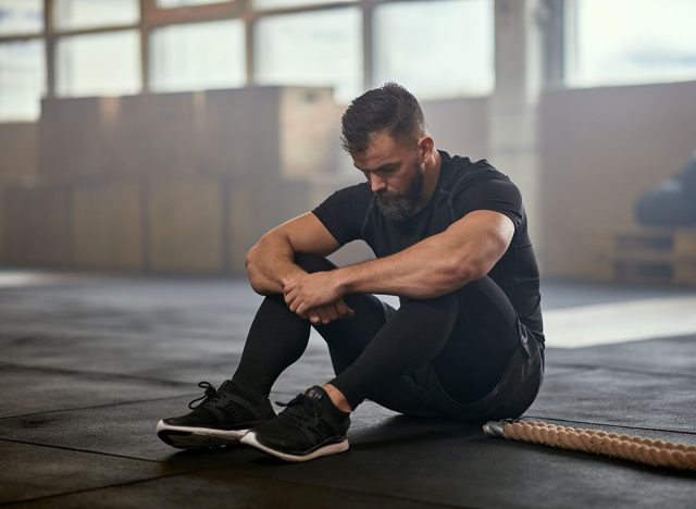 Fit young man in sportswear sitting on a gym floor and looking exhausted after a battle rope workout session
