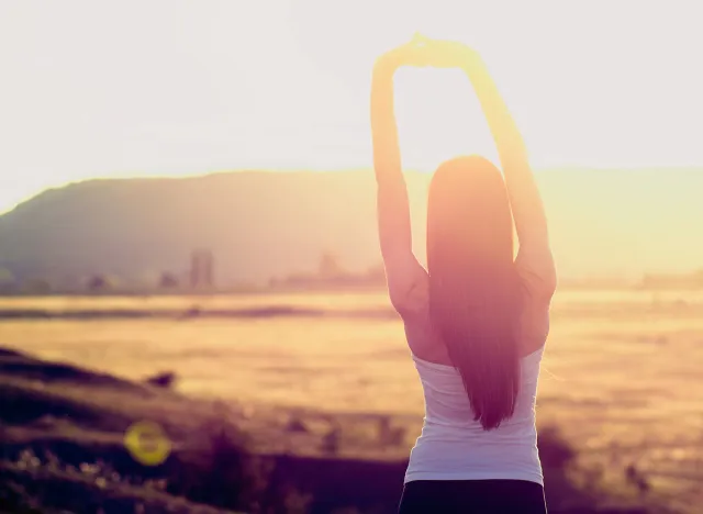 Happy celebrating winning success woman at sunset or sunrise standing elated with arms raised up above her head