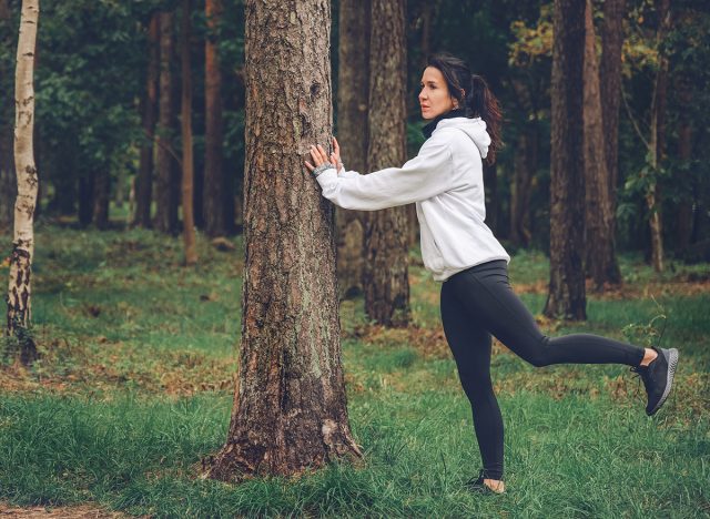 Young woman in white hoodie swinging leg in autumn park. Healthy exercising outdoors