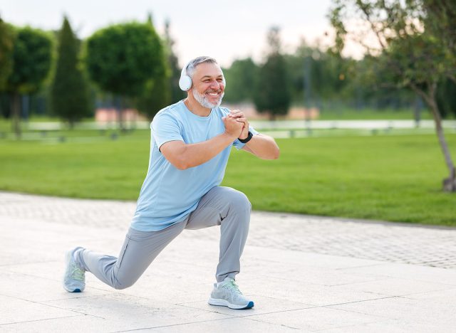 Full-length photo of happy smiling retired sportsman in sportswear and headphones doing front squat on one leg forward with hands folded outside in city park during routine workout in morning