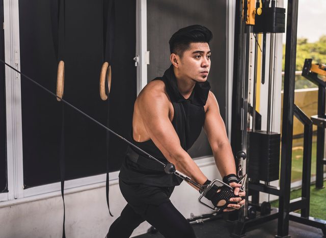 A fit and tanned asian man in his 20s does a set of cable crossovers at the gym. Wearing a black hooded low cut tank top. Training chest muscles in the gym.