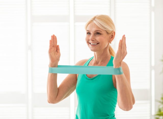 Active Lifestyle. Smiling Senior Lady Exercising With Resistance Band At Home, Portrait Of Sporty Mature Woman In Activewear Training With Fitness Equipment In Light Living Room Interior, Copy Space