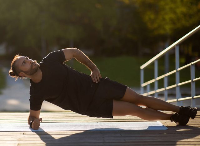 Outdoor Workout. Muscular guy in sportswear doing side elbow plank exercise working out on mat outside, full length shot. Training for strengthening core muscles, sport motivation