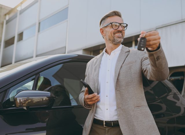 male buyer client wearing white shirt classic grey suit standing near black electric car while charging looking aside use mobile cell phone hold give keys outside. Business concept