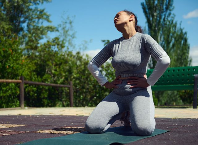 Young woman in activewear kneeling on a yoga mat in a park, appearing to have a stomach ache during a sunny outdoor workout session.