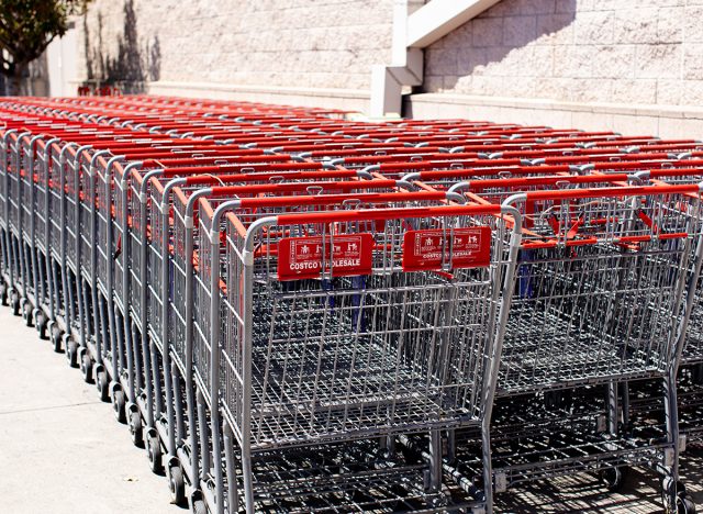 Rows of new metal Costco shopping carts with red handles outside Costco warehouse, with no customers in view, sunny day, against store wall