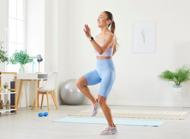 Happy beautiful sporty young Asian woman in sportswear doing a jumping and running in place exercise with high knee raises during her fitness workout in the living room at home