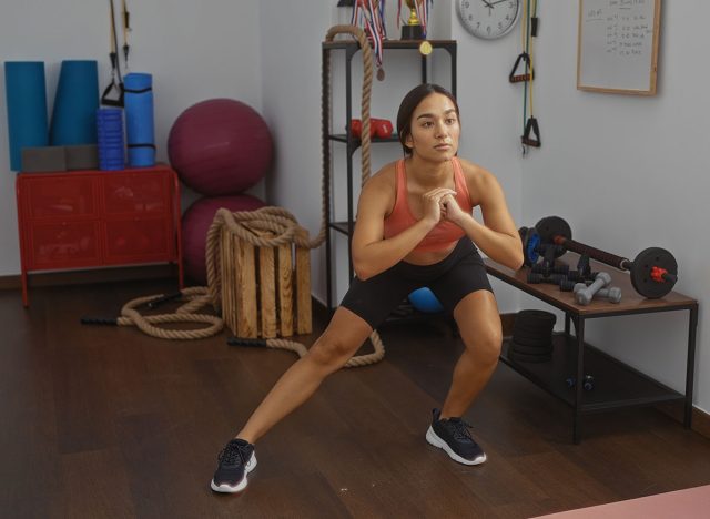 Woman exercising indoors wearing a workout outfit in a gym setting with various fitness equipment, demonstrating a focused lunge amid weights, ropes, and yoga mats in a home or fitness studio.