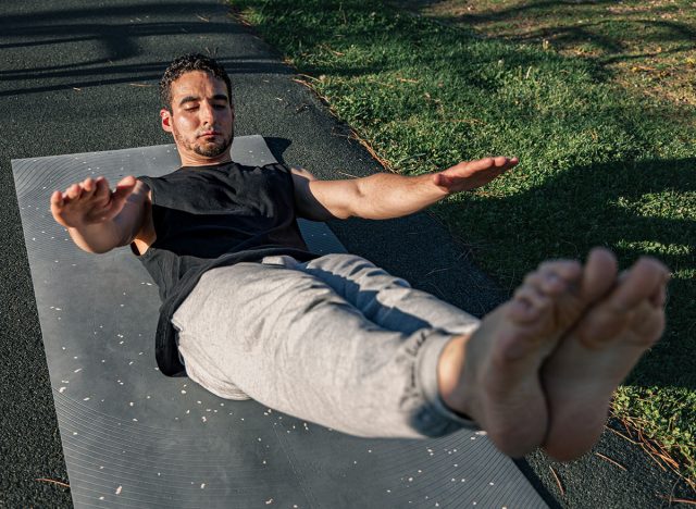 Young man performing hollow body hold on outdoor mat with arms extended and legs raised, showcasing core strength, balance, and control under sunlight