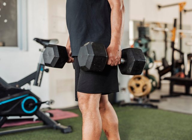 Anonymous man carries two dumbbells in a farmer's walk for forearm or grip strength and endurance training inside open-air gym.