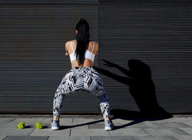 Back view athletic woman with perfect figure and buttocks doing squats against wall with copy space for your text message,fit female in sportswear squatting after training on black background outdoors