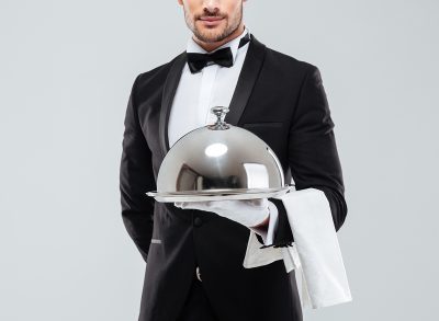 Waiter in tuxedo and gloves holding silver tray with lid and napkin